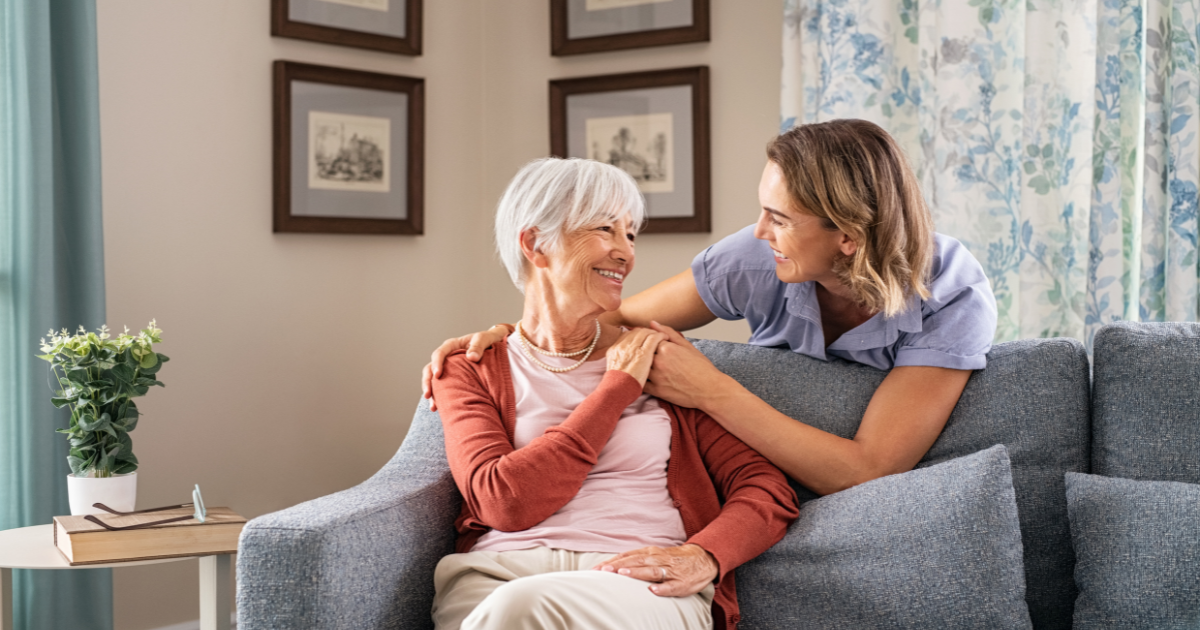 Smiling caregiver with hand over the shoulder of a happy elderly woman sitting on the couch.