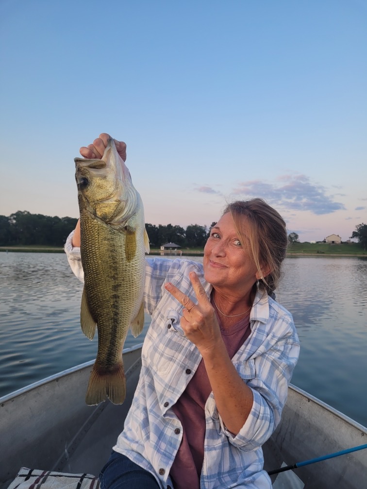 A smiling woman sits in a small boat on a calm lake at sunset, proudly holding up a large fish and flashing a peace sign.