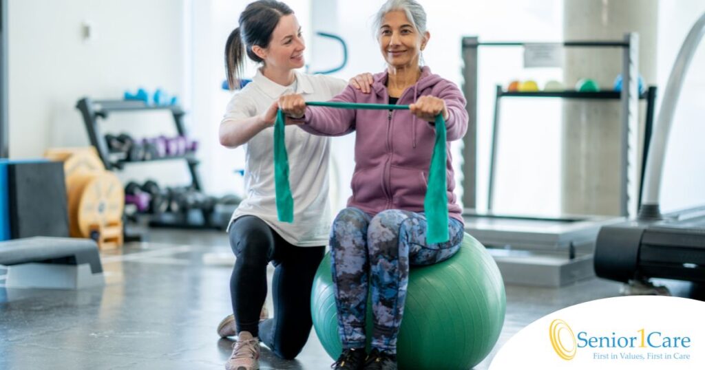 A care provider helps an older woman exercise with a resistance band and an exercise ball, representing how exercise can help with senior fall prevention.