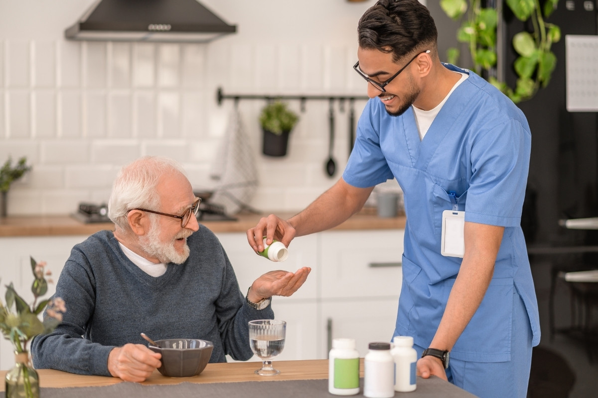 Warm and Dependable Medication Reminders in Carmel A friendly caregiver smiling and pouring a pill into the hand of an older gentleman at a bright kitchen table, representing trustworthy Medication Reminders in Carmel.