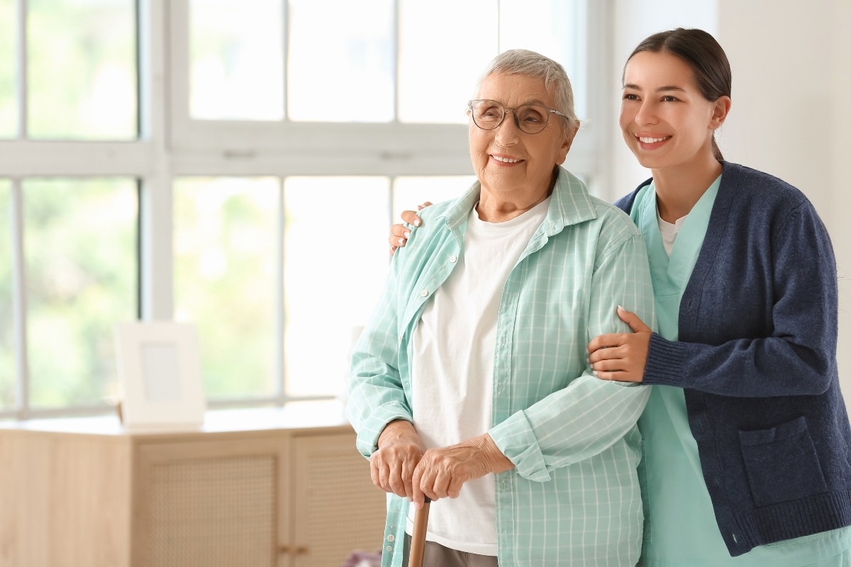 Compassionate and Supportive Personal Care in Carmel A cheerful caregiver gently supporting an older woman standing with a cane by a bright window, representing compassionate Personal Care in Carmel.