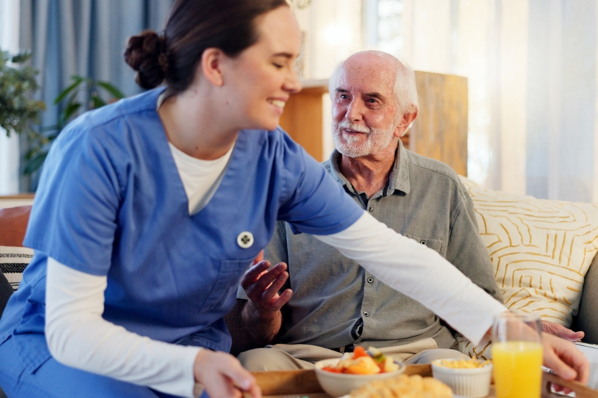 Compassionate In-Home Caregiver Providing Companionship Care in Carmel A smiling caregiver serving a nutritious meal to an older gentleman relaxing comfortably on a couch, representing Companionship Care in Carmel.